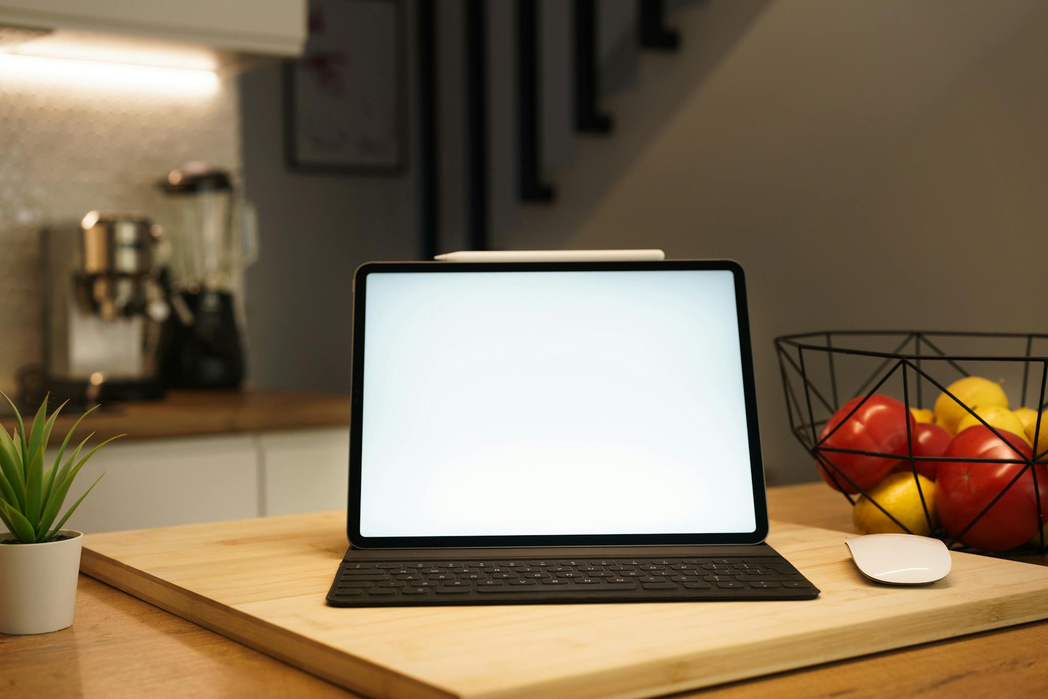 A modern kitchen workspace featuring a tablet with keyboard, mouse, and bowl of fruits on a wooden table.